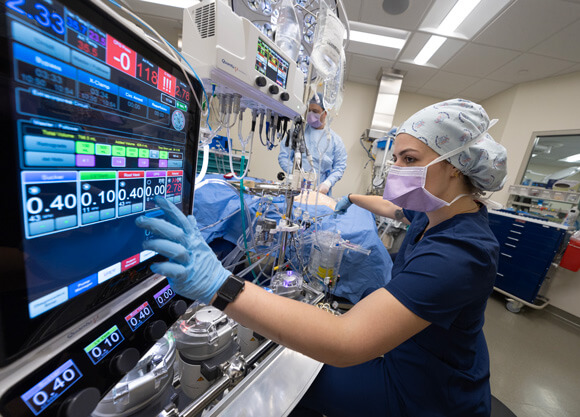 A student in a hair covering and mask clicking numbers on a screen in a simulated operating room.